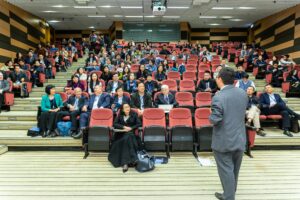 Professional setting of a business conference with attendees and a presenter in an auditorium.